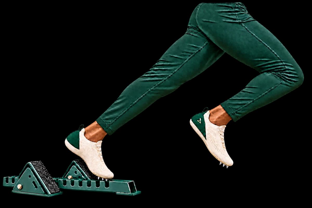Sprinter pushing from the left starting-block pedal with the right knee driving forward, wearing matching cream Cleats shoes